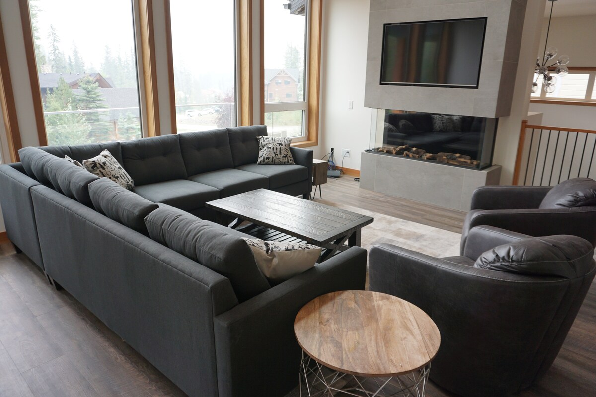 A spacious living area features a large sectional sofa arranged around a central coffee table. A modern fireplace is visible beneath a wall-mounted TV, and large windows allow natural light to fill the space, providing views of the surrounding landscape.