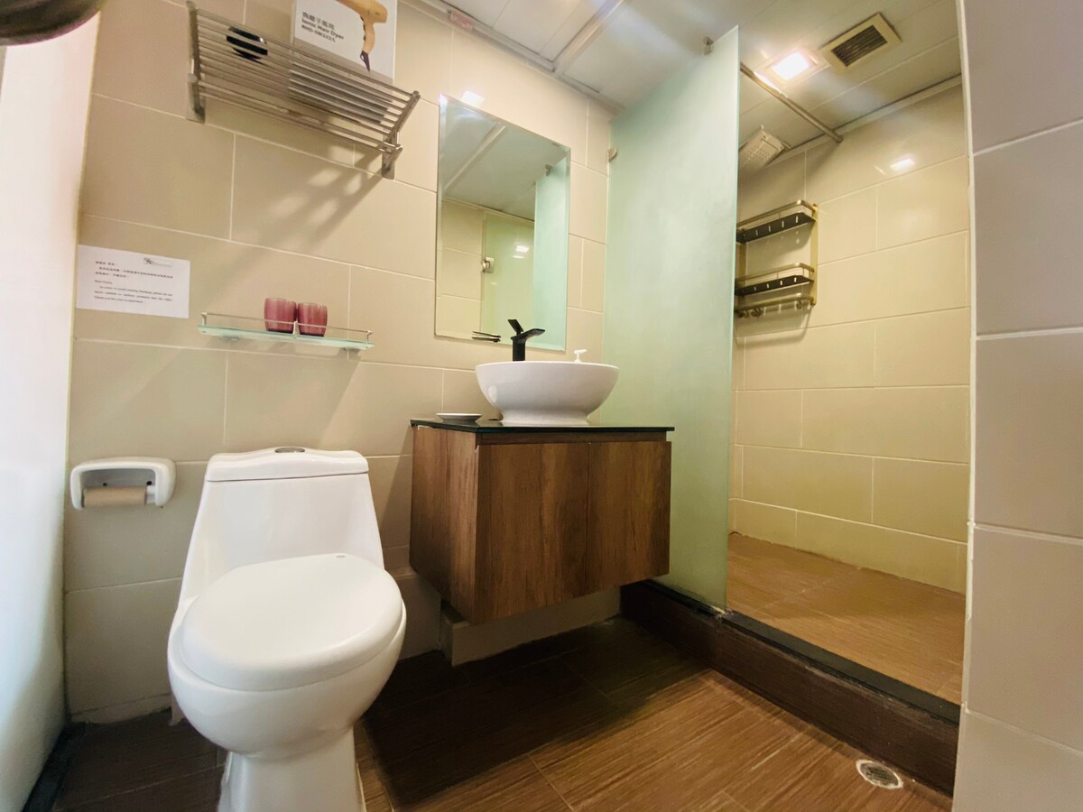 A modern bathroom features a sleek toilet and a compact wooden vanity with a vessel sink. A glass shower enclosure is visible, providing a light and airy feel. Shelving for toiletries is mounted on the wall, and neutral tiles complement the space.