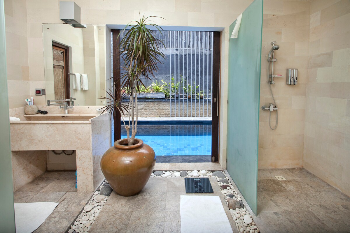 An airy modern bathroom includes a marble countertop with a sink and a large wooden mirror. Shower areas are separated by glass panels, featuring decorative plants. A large clay pot sits on the floor, which is adorned with small pebbles, and a glimpse of a pool is visible outside.