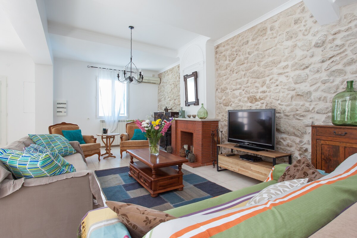 A spacious living area features a mix of natural stone walls and modern furnishings, including a comfortable sofa and armchairs. A wooden coffee table sits at the center, adorned with a vase of fresh flowers. A television is positioned nearby, complemented by decorative accents.