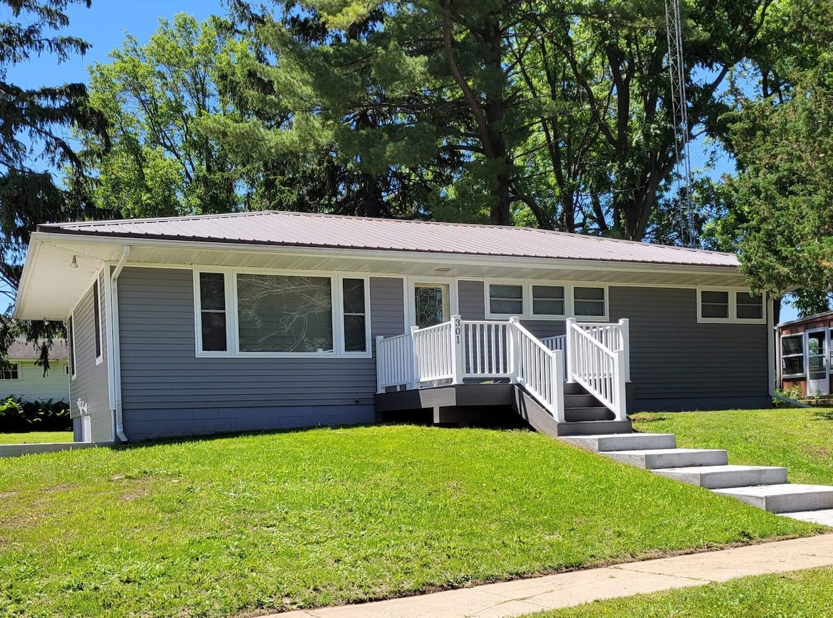 A modern single-story home is set on a manicured lawn, featuring a grey exterior and multiple large windows. A wooden ramp leads to a welcoming front porch, with several steps leading up to the entrance. Lush trees provide natural shade in the surrounding area.