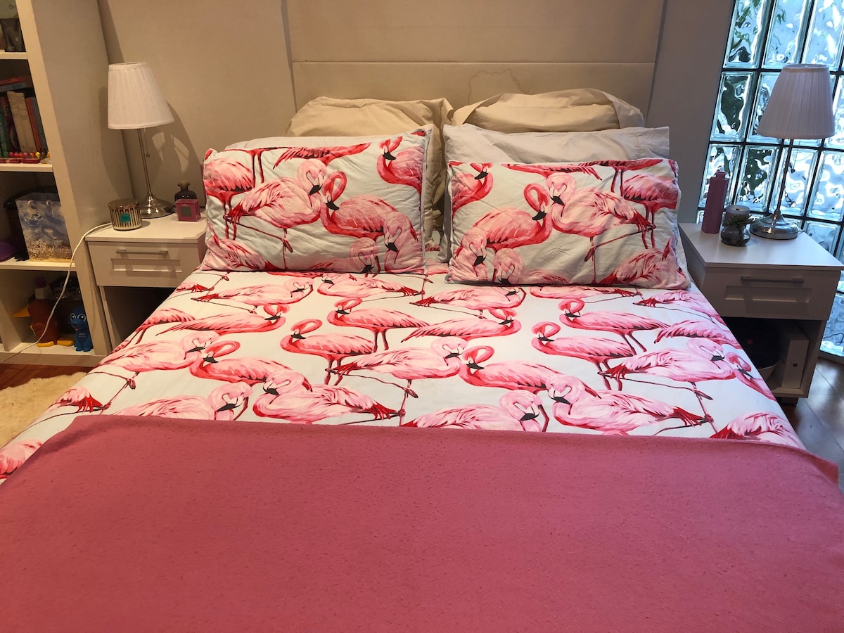 A spacious bed is adorned with bright pink flamingo-patterned bedding. Two white bedside lamps are positioned on either side, providing subtle lighting. A light pink throw blanket adds a cozy touch to the bed, which is set against a soft grey headboard.