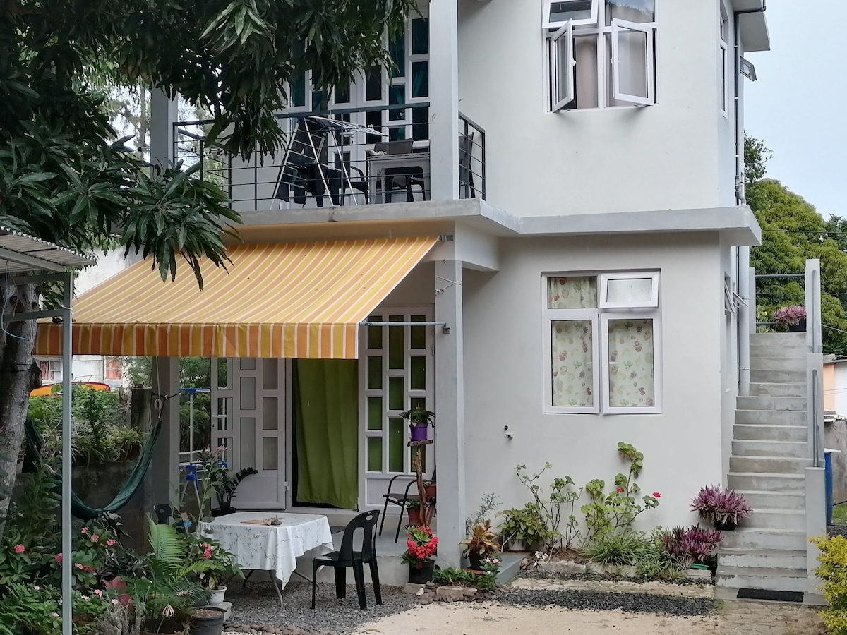 A two-story building is showcased with a striped awning shading a small outdoor seating area, including a table and chair. Surrounding greenery and flowering plants enhance the entrance, while steps lead to an upper level. Large windows allow natural light into the interior.