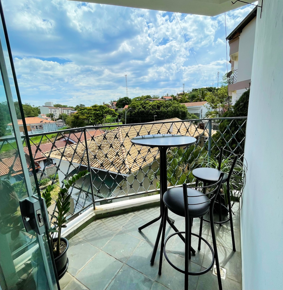 A balcony is visible, featuring a round table accompanied by two high-top chairs. A view of the surrounding neighborhood can be seen through the glass doors, with greenery in the foreground and buildings in the background.