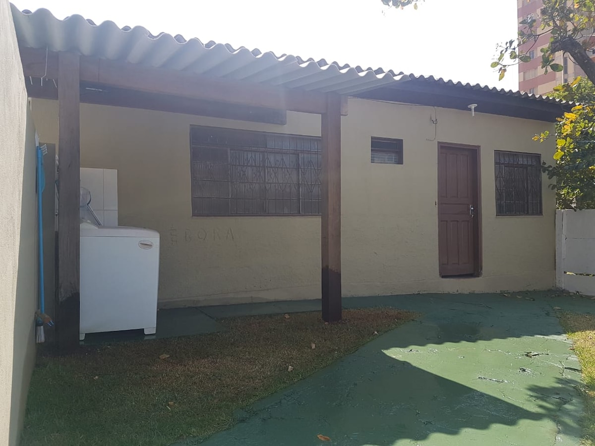 An exterior view of the house showcases a covered porch with a wooden roof. A washing machine is positioned on the side, with grass and green concrete surrounding the area. Windows allow natural light to enter the interior spaces.