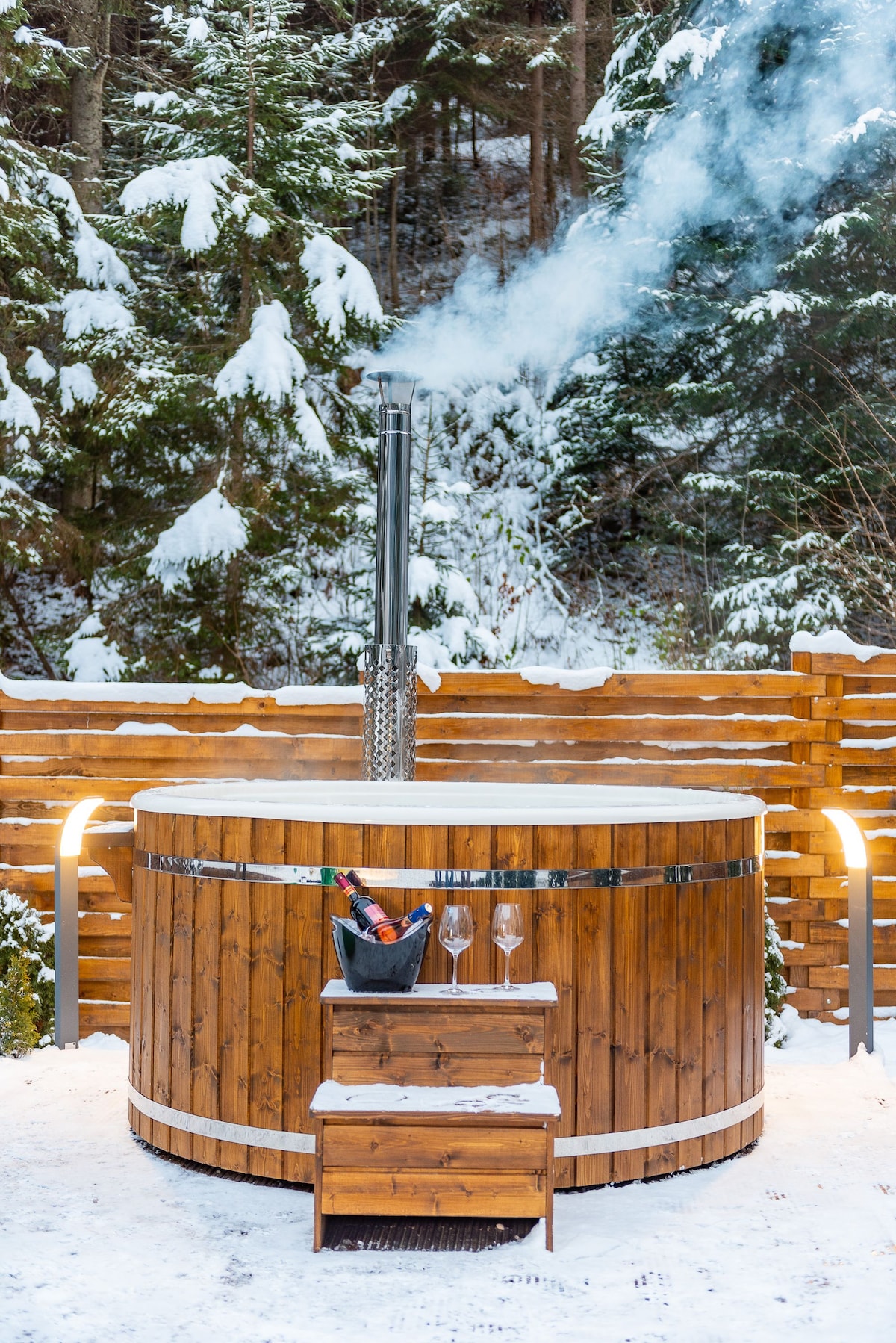 A wooden hot tub is positioned outdoors, surrounded by snow-covered trees. Smoke is gently rising from a chimney. A small wooden step leads up to the tub, which is accompanied by a bucket of ice and two glasses, suggesting a relaxing atmosphere.