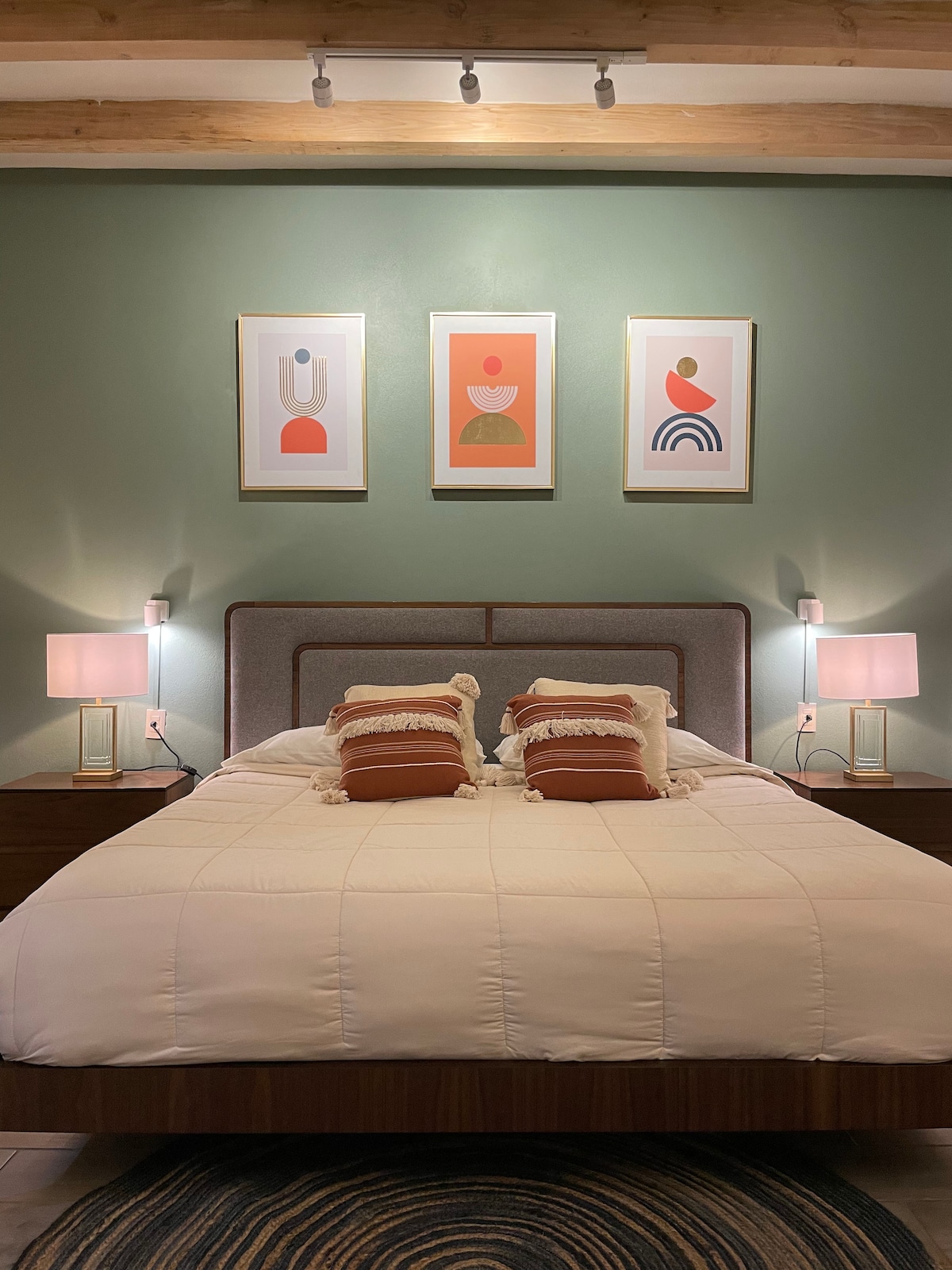 A well-lit bedroom features a spacious bed with a soft white comforter, accented by patterned throw pillows. Two bedside lamps provide gentle illumination beside the bed. Above, three framed art pieces with abstract designs add a touch of color to the green walls.