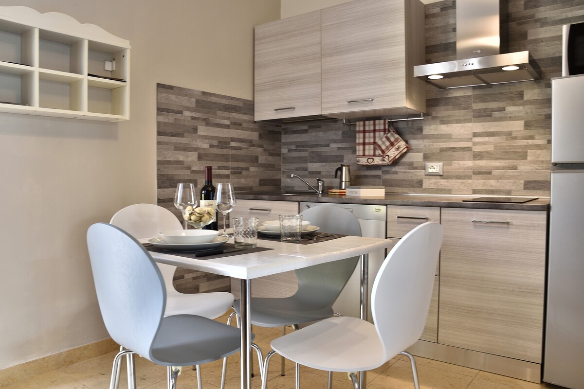 A modern kitchen area is highlighted by gray cabinets and a minimalist white table set for four. Glassware and dinner plates are arranged on the table, while contemporary appliances, including a stovetop and sink, provide practical amenities for cooking.