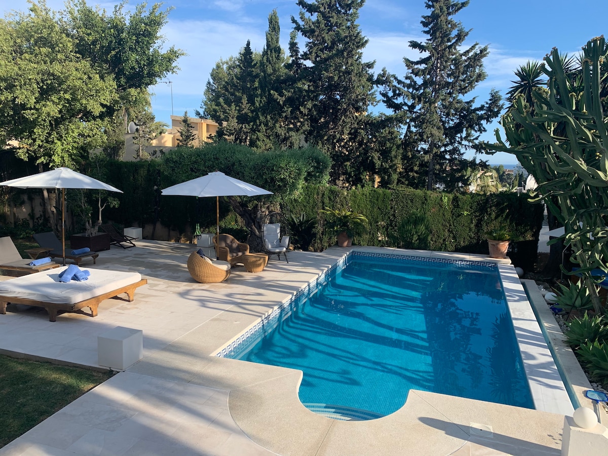 A serene garden features a saltwater swimming pool with a clear blue sheen, surrounded by lounge chairs and umbrellas. Lush greenery provides privacy, while decorative pots anchor the space, creating a peaceful area for relaxation and enjoyment.
