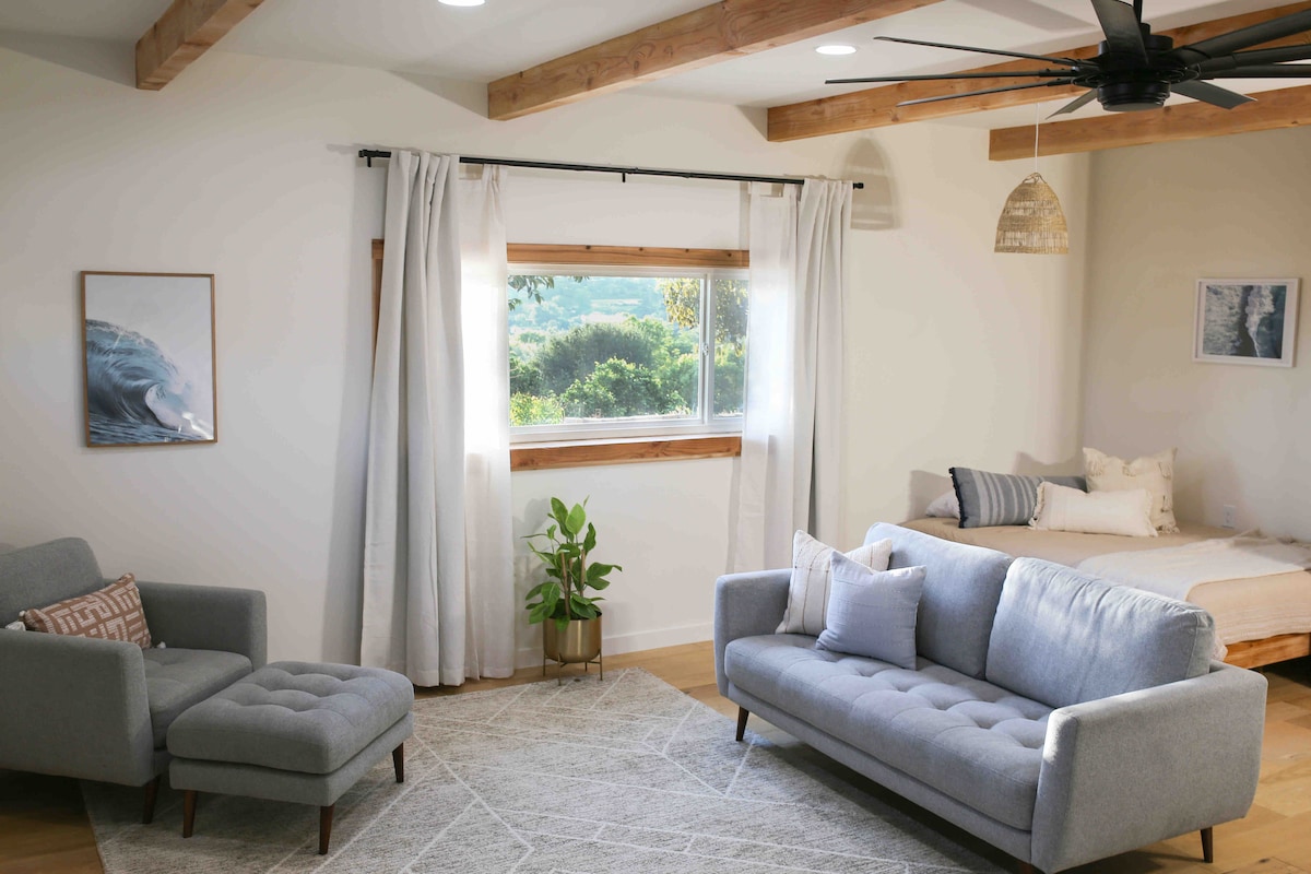 A modern living area features a light gray sofa and a matching armchair, positioned across from a spacious bed. Natural light enters through a window adorned with white curtains, offering views of the surrounding greenery. Textured area rug complements the warm wood accents on the ceiling.