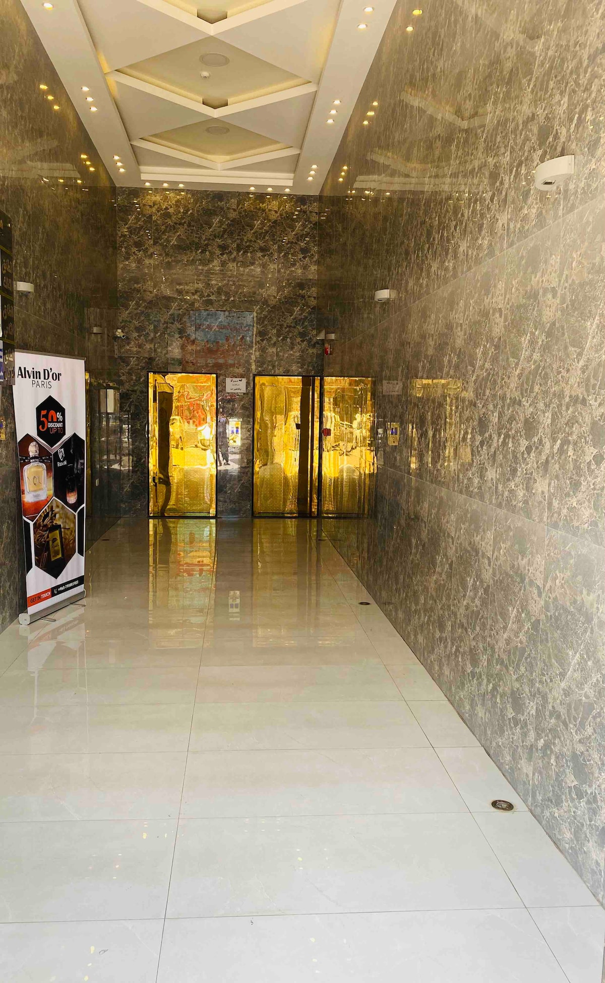 A modern hallway is characterized by polished marble walls and a glossy tiled floor. Golden double doors leading to the elevators are visible at the end, framed by soft lighting. A promotional banner is positioned to the left of the entrance, adding a touch of color.