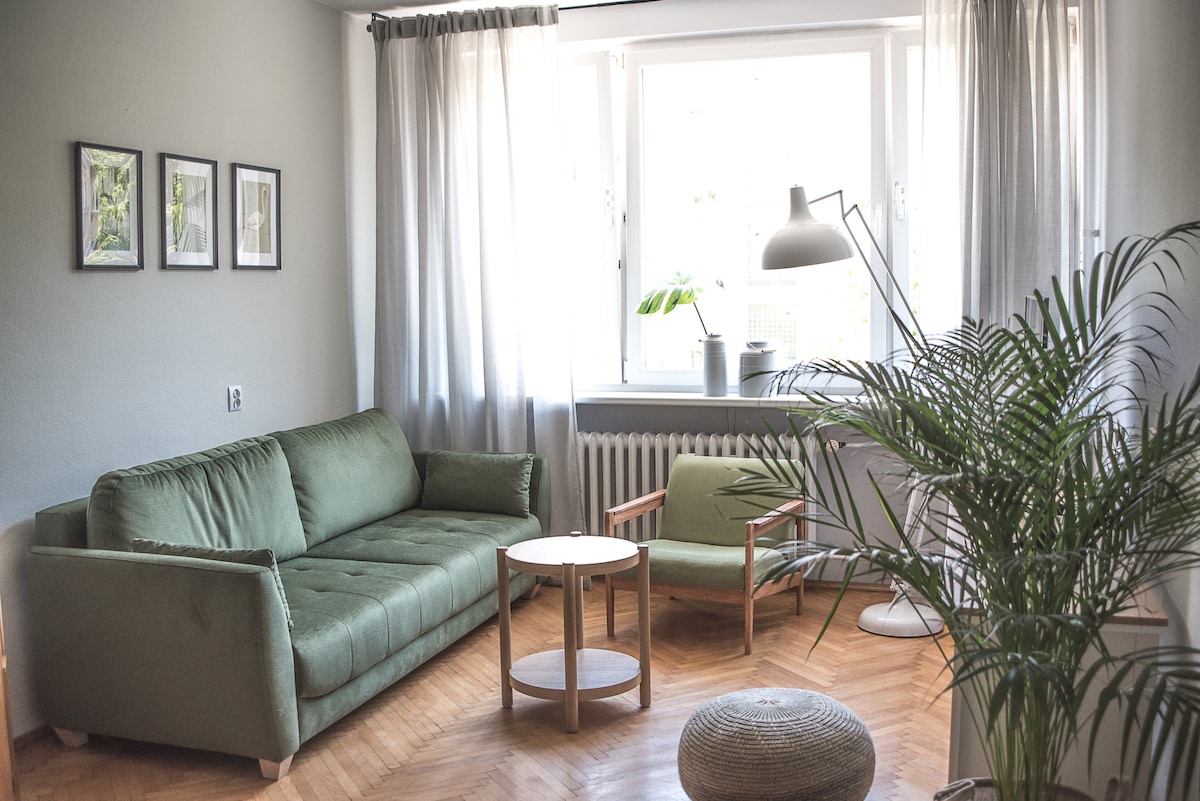 A comfortable living area features a green sofa and an accent chair, complemented by a round wooden table. Natural light floods through the large window, adorned with light curtains. A potted plant adds a touch of greenery, enhancing the overall ambiance.