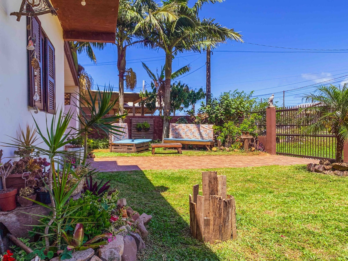 A spacious patio area is surrounded by greenery, featuring two sun loungers and decorative plants. The space is embraced by a brick wall and an electronic gate, providing a safe environment. A rustic seating area made of logs adds a natural touch.
