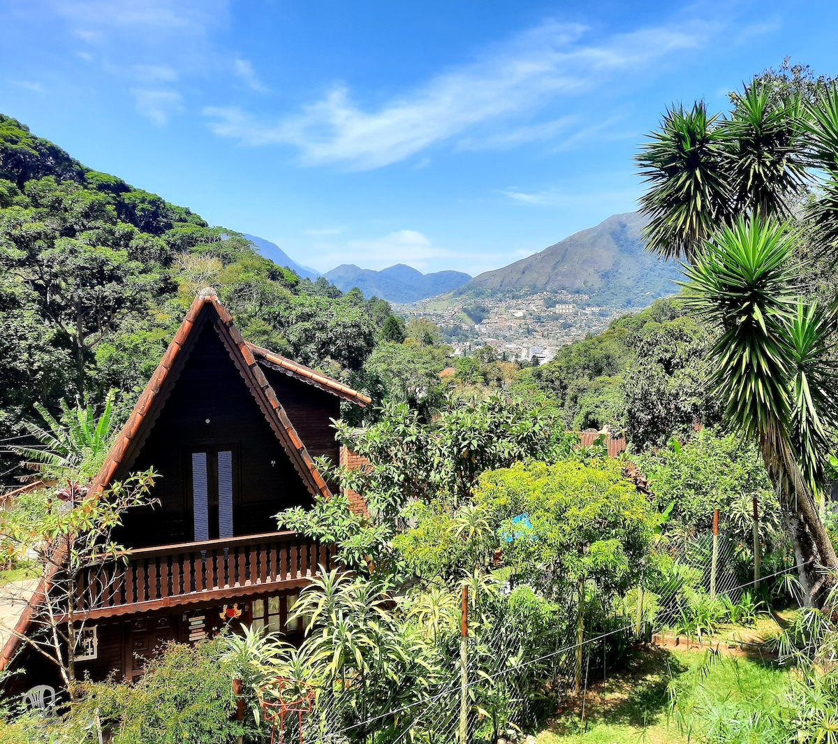 A two-story wooden chalet is nestled among lush greenery, showcasing a sloped roof and balcony. The landscape features rolling hills and distant mountains under a clear blue sky, while a variety of trees and plants surround the property, creating a serene atmosphere.