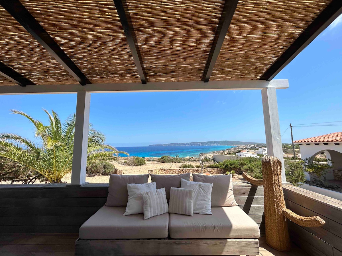An outdoor seating area features a spacious couch adorned with light-colored cushions. A natural fiber roof provides shade, while the view showcases the coastline and distant hills, lending a serene backdrop to the setting.