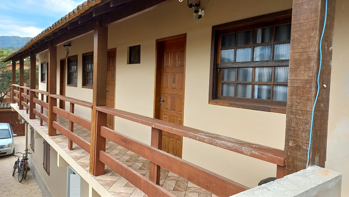 A neutral-colored building exterior features several wooden doors along a covered balcony. Wooden railings provide a simple barrier, while a tiled floor adds texture. Surrounding greenery and mountains are visible in the background, contributing to a peaceful and natural setting.