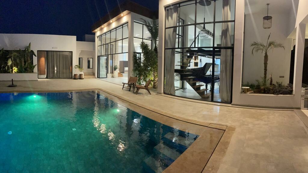 An outdoor view showcases an illuminated pool reflecting soft turquoise hues. The villa's expansive glass windows reveal a modern interior, while an elegant staircase and greenery are visible. Subtle lighting enhances the tranquil ambiance of the evening setting.