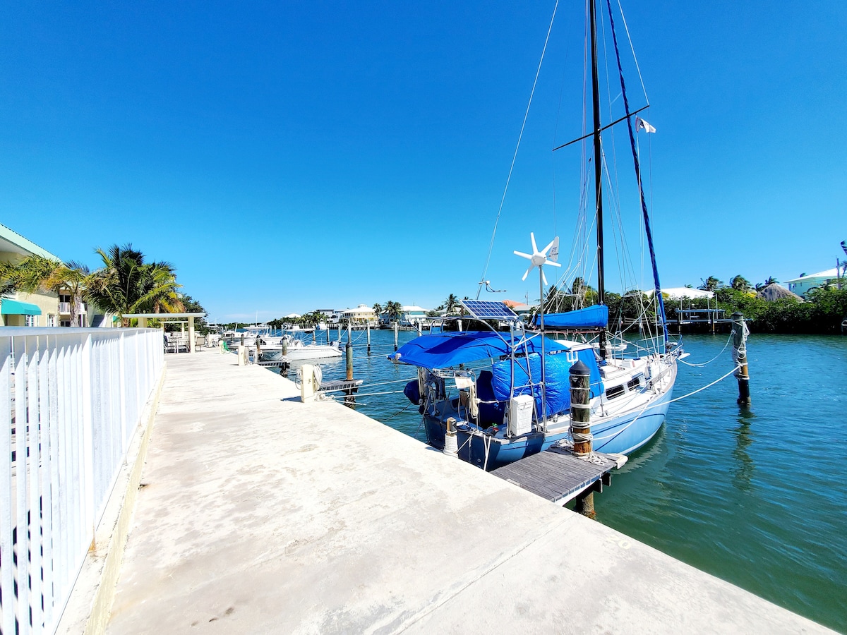 The image captures a serene waterfront scene featuring a calm waterway with moored boats. A concrete dock extends into the water, flanked by a white railing. A sailboat with a blue cover is prominently positioned, surrounded by lush greenery in the background.