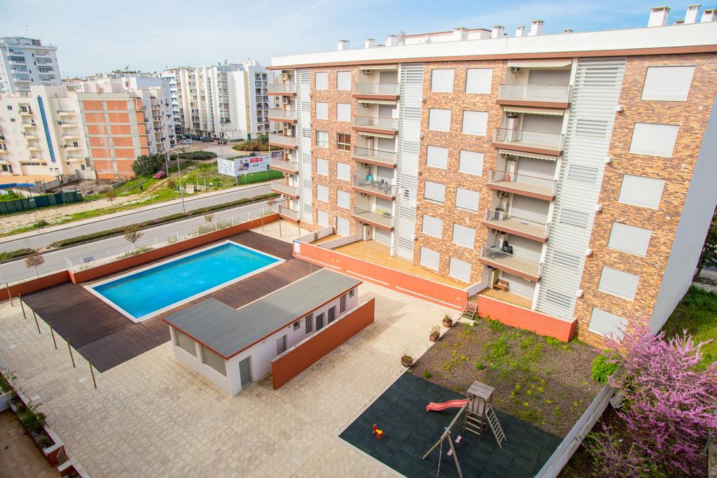 The image captures an overview of the apartment complex with a swimming pool in the foreground. Surrounding the pool, sun loungers and a wooden deck are visible, while a children's play structure is also present. The building features several balconies and mixed brick facades.
