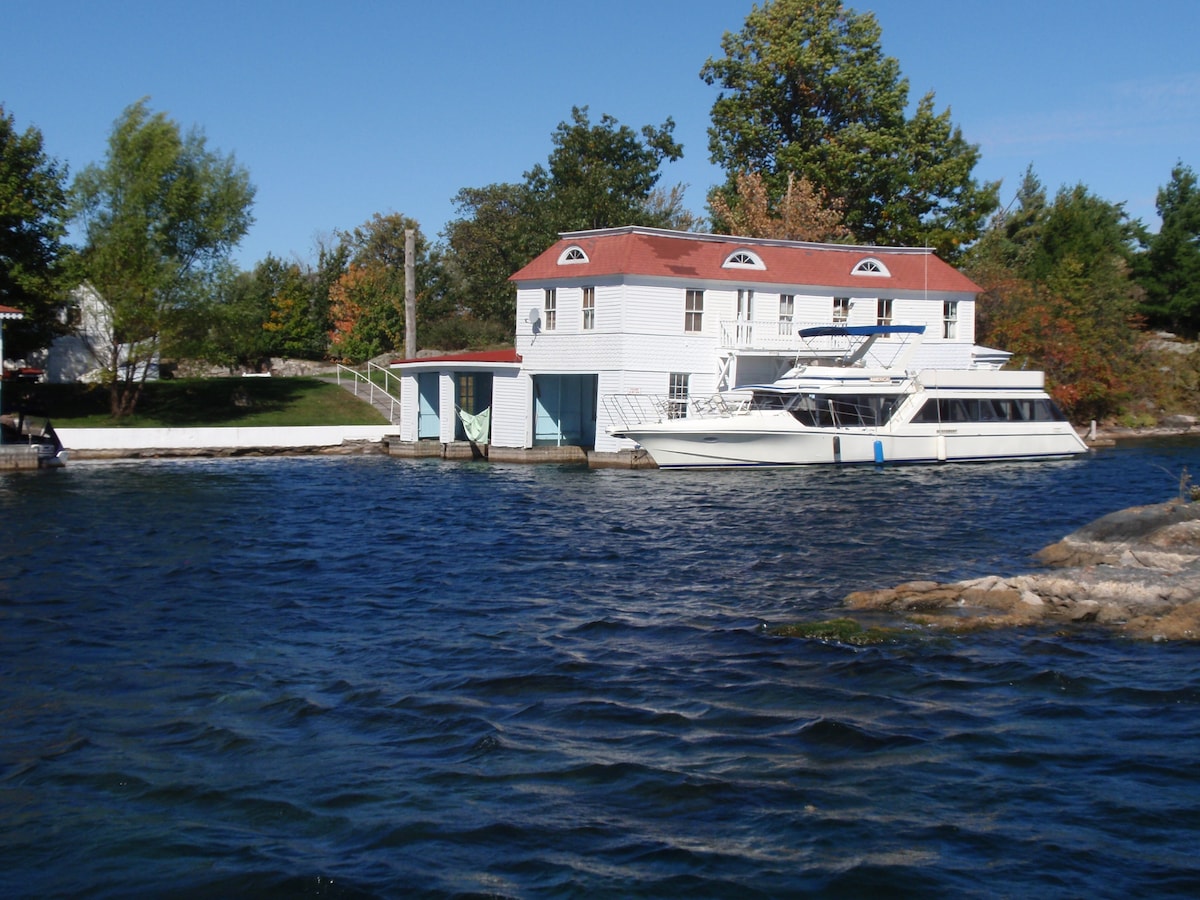 A charming two-story structure is visible, featuring a white exterior and red roof. A private boat is docked nearby, with calm water surrounding the property. Lush greenery and trees are present in the background, adding to the scenic riverfront setting.