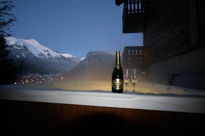 An outdoor jacuzzi is set against a mountain backdrop, illuminated by soft evening light. A bottle of champagne and two glasses are positioned at the edge, suggesting a relaxing experience. The distant village lights twinkle in the valley below.