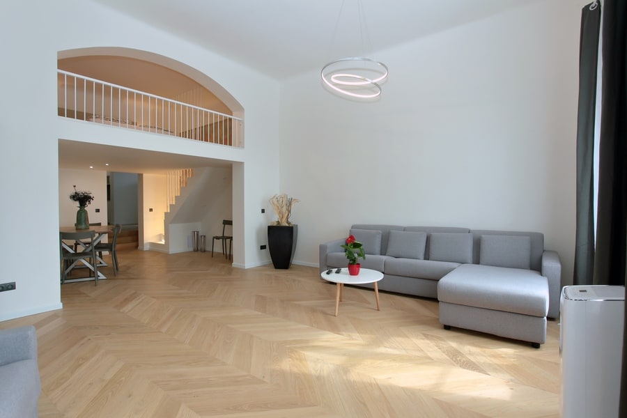A spacious living area showcases light-colored wooden flooring and modern furniture. A gray sectional sofa is positioned near a minimalist coffee table, with a potted plant adding a touch of greenery. An upper loft area is visible, enhancing the open ambiance of the room.