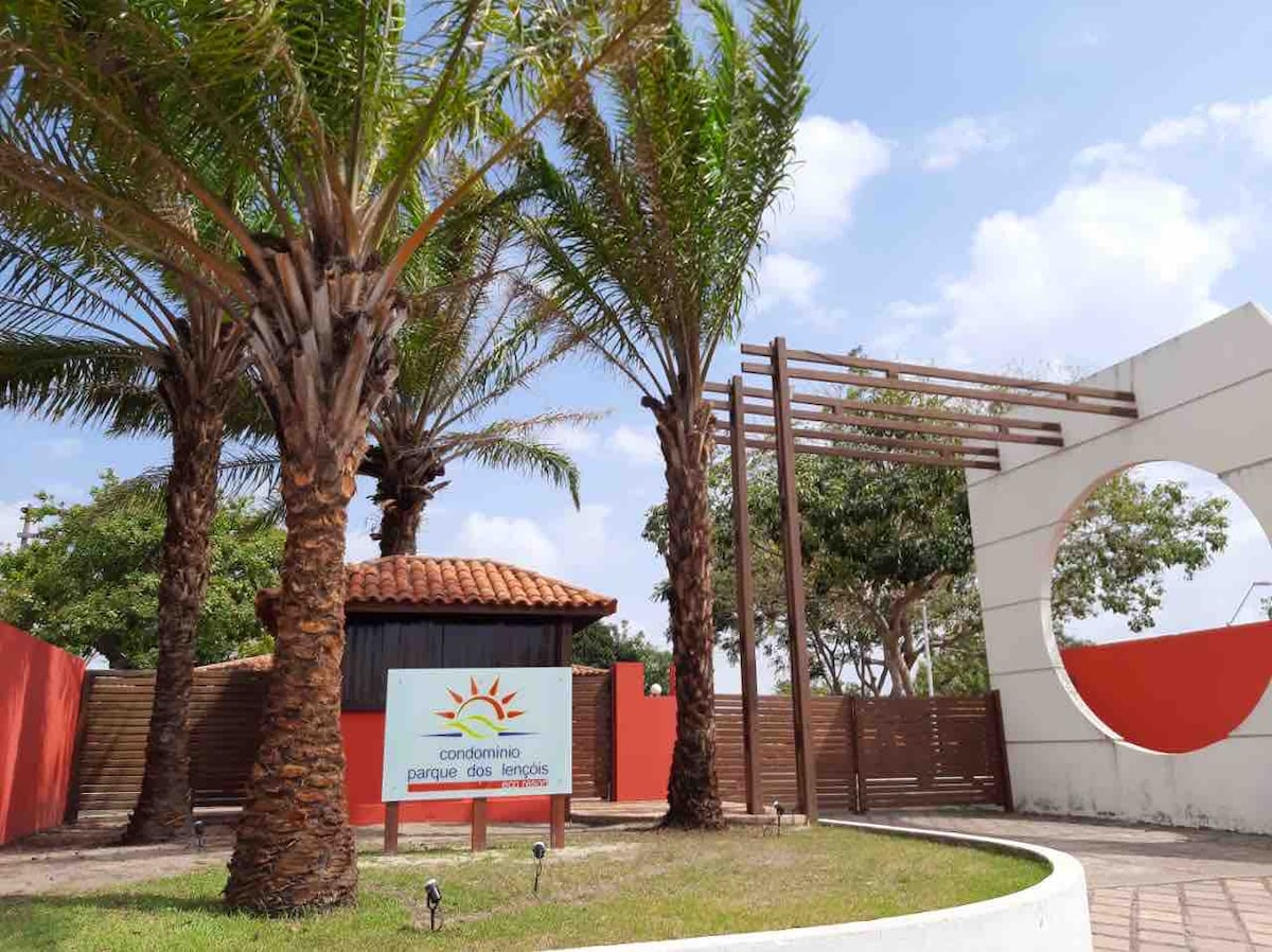 The entrance to Parque dos Lençóis Eco Resort is framed by palm trees and features a welcoming sign. The design combines wooden elements and a circular cutout in the wall, leading into a well-maintained landscape under a clear blue sky.