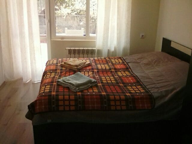 A sleeping area is featured, with a spacious bed covered by a patterned blanket. Neatly folded towels are placed on top. Natural light enters through large windows dressed in sheer curtains, illuminating the wooden flooring.