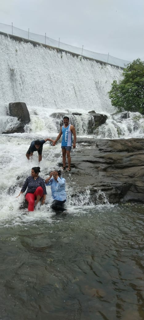 A group of individuals is seen wading in shallow water near a rocky area, with a dam cascading water in the background. Smooth stones are scattered across the foreground, and lush greenery is visible along the edges of the scene.