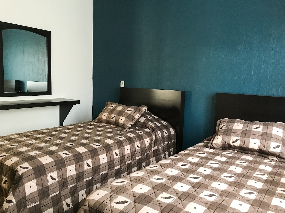The sleeping area features two spacious beds with patterned bedding, complemented by a dark accent wall. A mirror is mounted above a small shelf, adding practicality to the space.