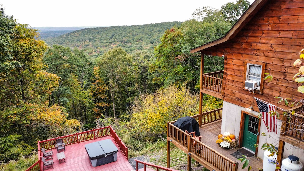 A rustic log cabin is set against a backdrop of rolling hills covered in changing foliage. A spacious deck features a hot tub, seating arrangements, and a charcoal grill. The natural landscape provides a serene view of the surrounding mountains.