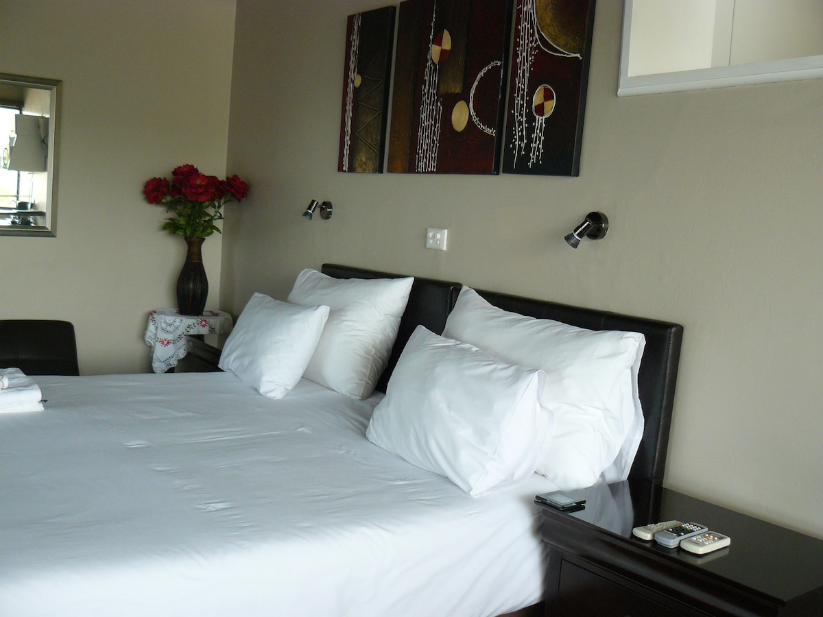 A sleeping area is featured with a spacious bed dressed in crisp white linens. Soft pillows are arranged neatly at the headboard. A decorative wall art piece adds color above the bed, while a small vase of flowers brings a touch of nature to the bedside.
