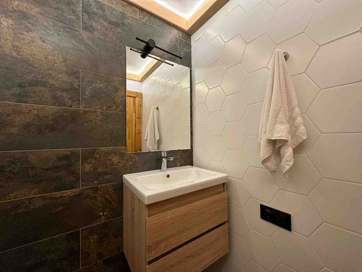 A modern bathroom features hexagonal tiles in soft tones, complemented by a wooden vanity with two drawers. A sleek mirror is mounted above a white sink, with a towel hanging nearby. Warm lighting enhances the contemporary design.