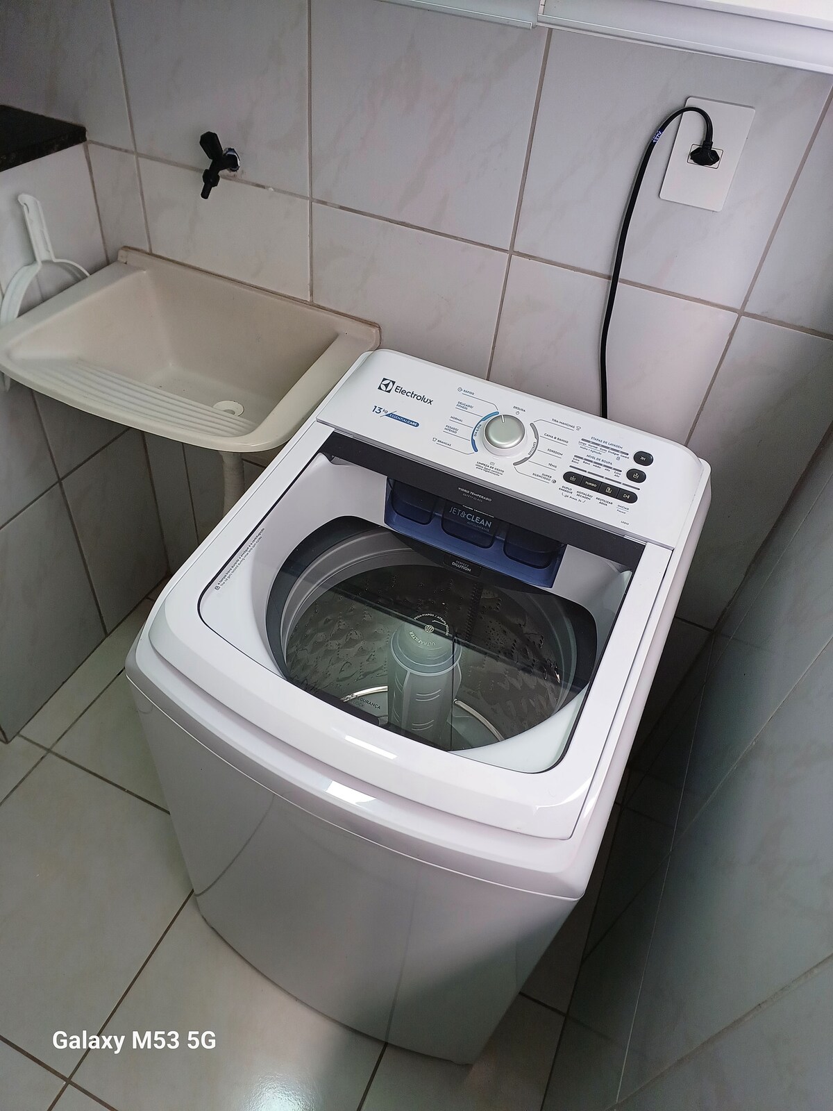 A washing machine is displayed prominently in a tiled laundry area. The machine features a clear lid, controls at the top, and is positioned next to a sink. A power outlet is visible in the background, ensuring convenient access for use.