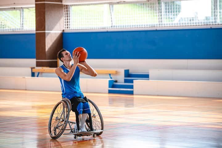 A person in a wheelchair wearing a blue jersey is poised to shoot a basketball in an indoor court.