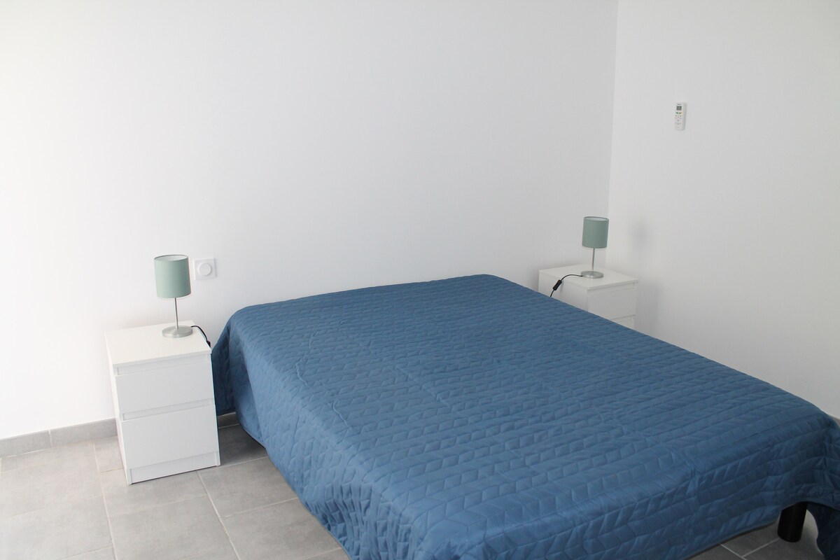 A spacious bedroom is presented with a large, neatly made bed covered in a blue quilt. Two matching bedside tables with lamps are placed on either side, and light grey tiles cover the floor, contributing to a clean and minimal aesthetic.