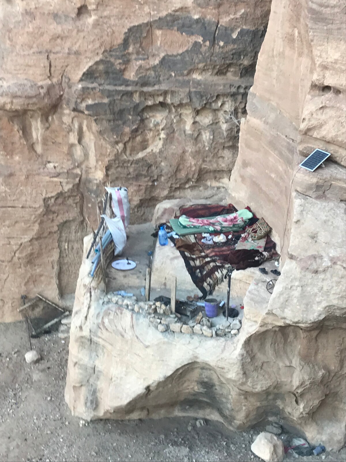 A cozy outdoor lounge area is set against a rocky backdrop, featuring colorful blankets and rugs arranged on the ground. A small solar panel is visible nearby, and various personal items are scattered around, reflecting a simple, natural living space.