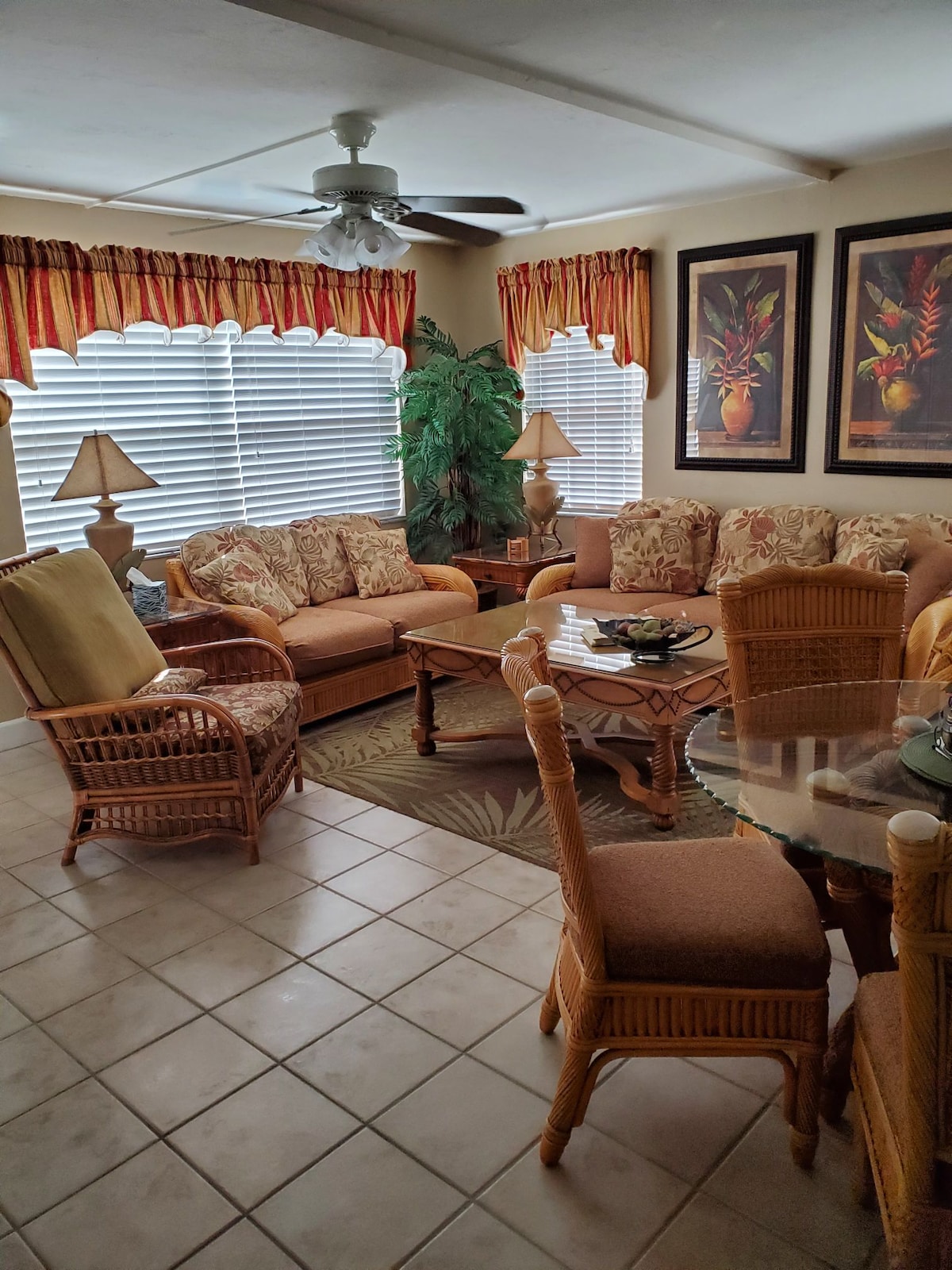 A comfortable living area is showcased, featuring a corner sofa and armchair with floral patterns. A round table sits in the center, surrounded by four upholstered chairs. Natural light filters through the white blinds, illuminating the space adorned with plants and framed artwork.