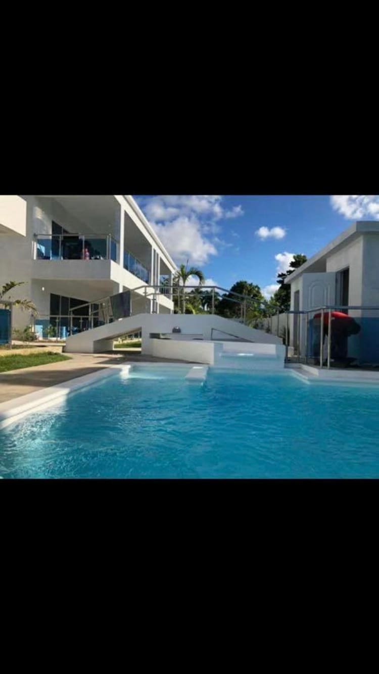A clear blue swimming pool is depicted, reflecting the sunlight. Modern white structures are visible on either side, with landscaped greenery surrounding the area. A staircase leads from the pool area to a terrace, enhancing accessibility and connection between indoor and outdoor spaces.