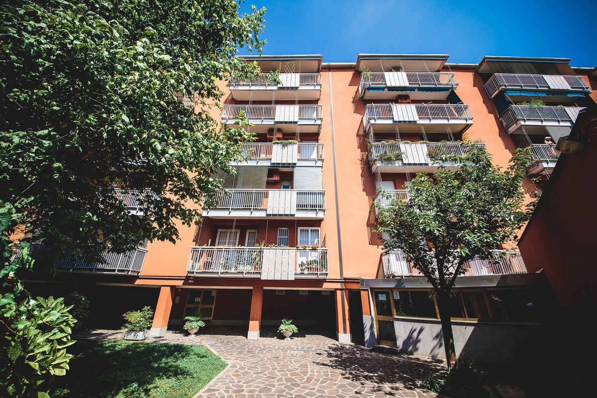 The residential building features a vibrant orange exterior. Multiple balconies with greenery are visible on each floor, complemented by trees and landscaped areas at the base. A stone pathway leads to the entrance, enhancing the inviting atmosphere of the property.