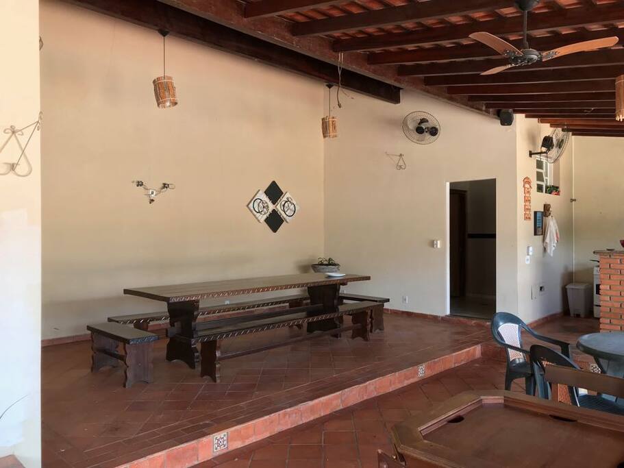 área de lazer coberta com mesa grande para refeições, ventiladores, banheiros feminino e masculino na área próximo a churrasqueira e piscina.
