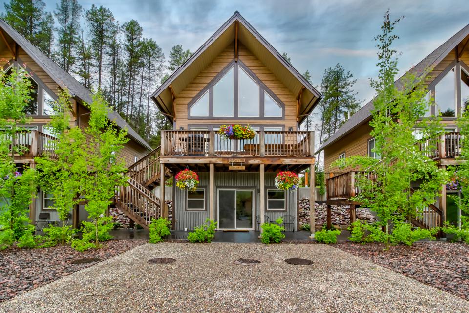 The chalet features a modern design with a triangular roof and large windows. Flower baskets adorn the balconies, and lush greenery surrounds the entrance. A stone pathway leads to the front door, enhancing the natural aesthetics of the setting.