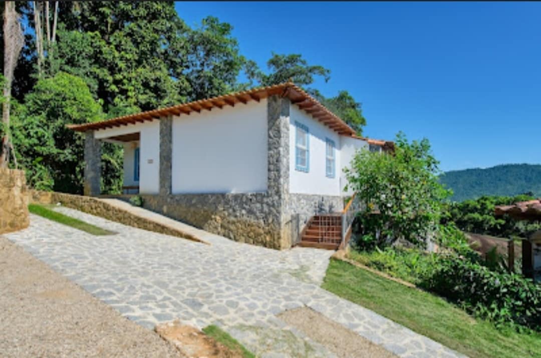 A single-story house is positioned on a gently sloping pathway, featuring a stone facade and a wooden roof. Large windows provide views of the lush greenery surrounding the property. Steps lead up to the entrance, enhancing accessibility to this tranquil setting.