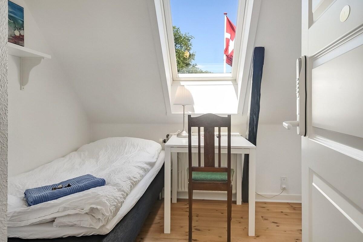 A simple room is equipped with a bed and a small desk with a chair. Natural light is filtered through a skylight above. A lamp sits on the desk, and a folded towel is placed on the bed. A flag is visible outside through the window.