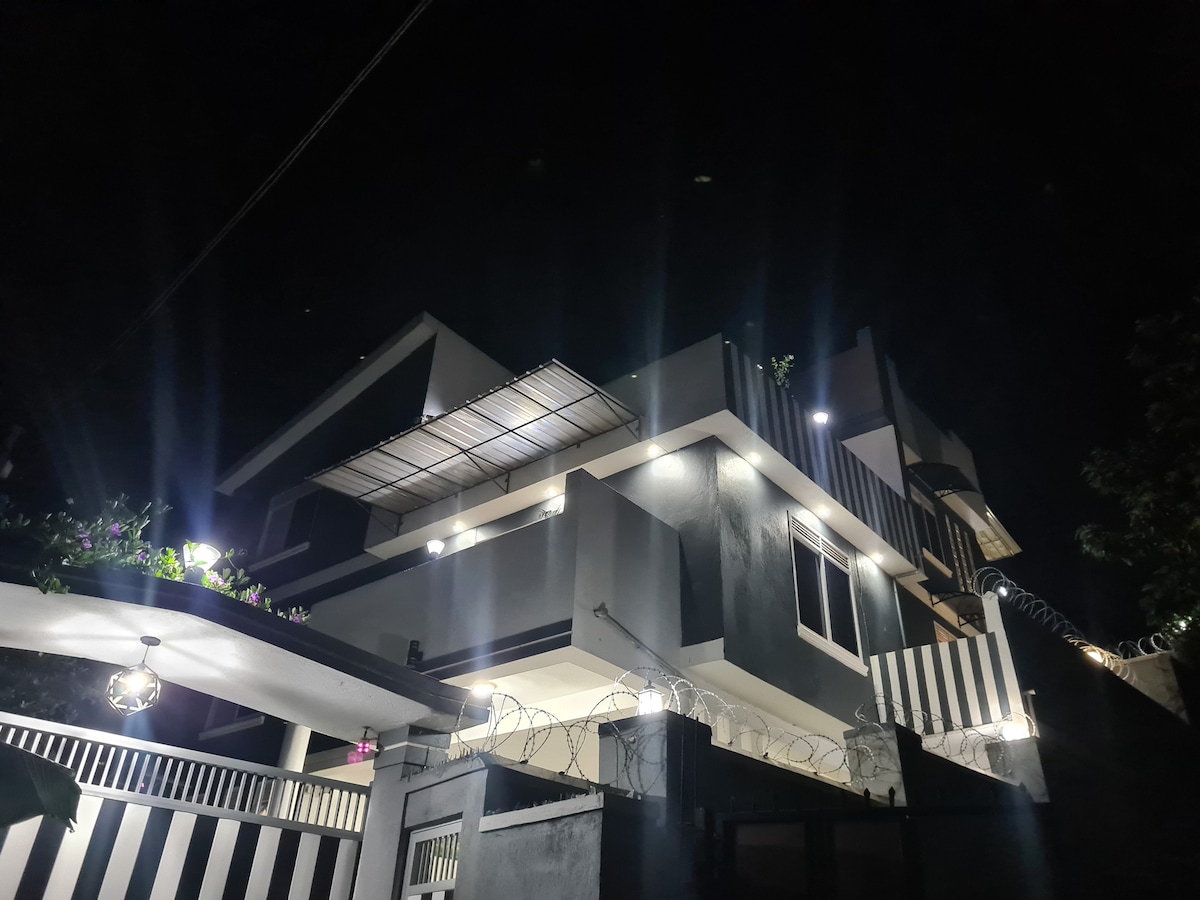The contemporary property is illuminated at night, highlighting its modern architecture. Soft lights accentuate the exterior features, and a balcony extends from the upper level. A secure entrance is visible at the front, surrounded by decorative fencing.