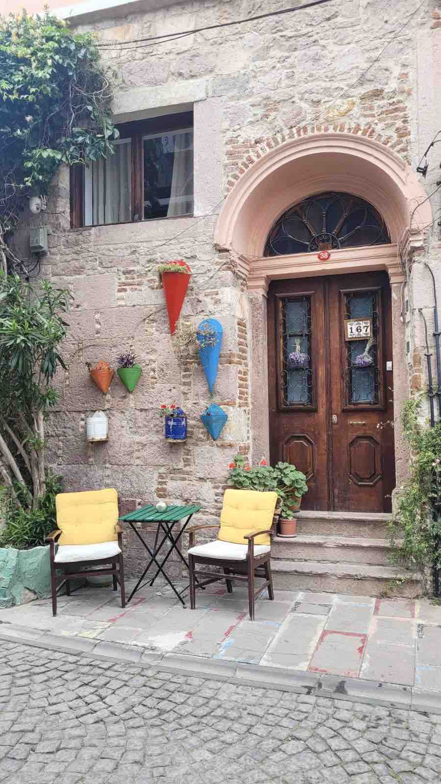 The charming entrance to a historic building features a set of wooden double doors with glass panels. Colorful planters adorn the walls, while two yellow chairs and a small table create a welcoming outdoor seating area beside a textured stone pathway.