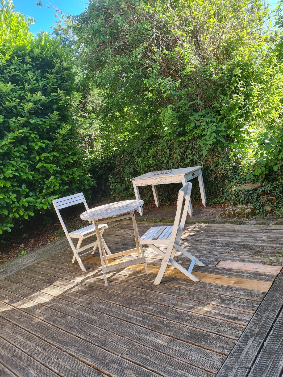 A small outdoor area is highlighted, featuring a round wooden table accompanied by two white folding chairs. Lush greenery surrounds the space, providing a sense of privacy and a natural setting. Sunlight filters through the leaves, casting soft shadows on the wooden deck.