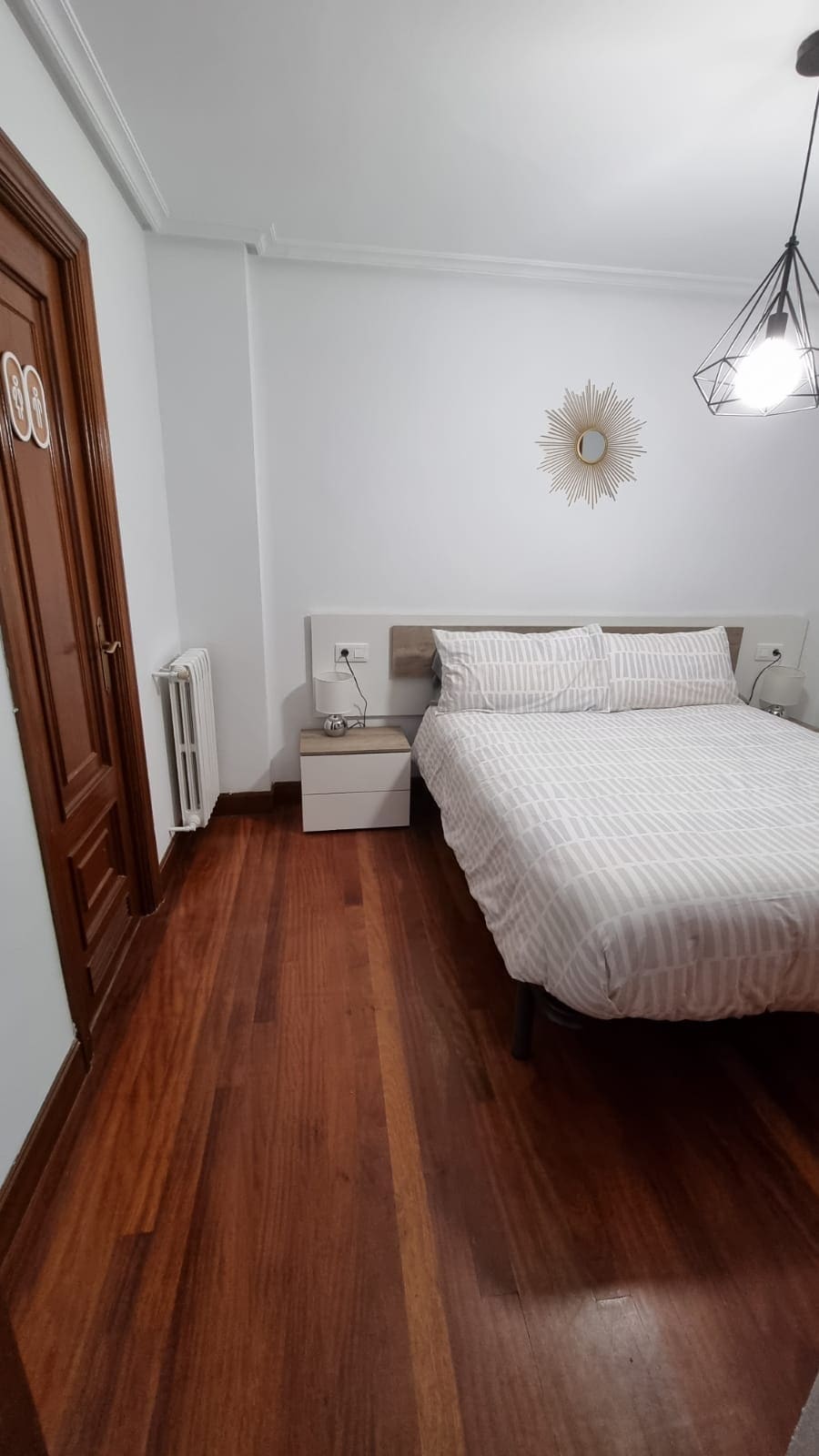 A cozy bedroom is presented with a spacious bed featuring light-colored linens. A bedside table is positioned beside the bed, and a mirror hangs on the wall. The warm wooden flooring contrasts with the white walls, and natural light subtly illuminates the space.