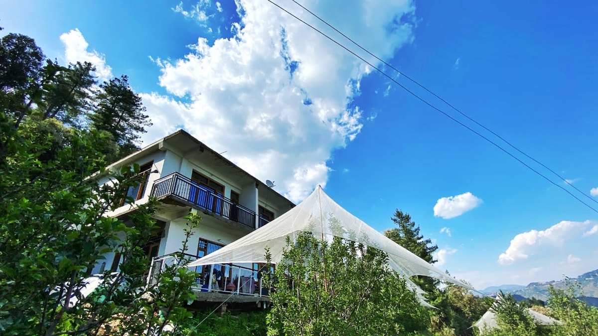 A two-story building is positioned amidst lush greenery, featuring a balcony with a contemporary awning. The clear blue sky, adorned with fluffy white clouds, provides a serene backdrop. Surrounding apple trees contribute to the natural ambiance, enhancing the connection with the outdoor landscape.