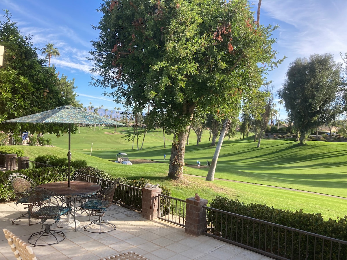 An outdoor patio area features a round dining table surrounded by metal chairs, shaded by an umbrella. Lush greenery and a tree provide a natural backdrop, while expansive views of the golf course and mountains stretch into the distance.